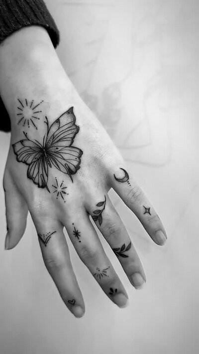 Fine line butterfly tattoo with star accents on the back of a hand, by Fawn Harper.